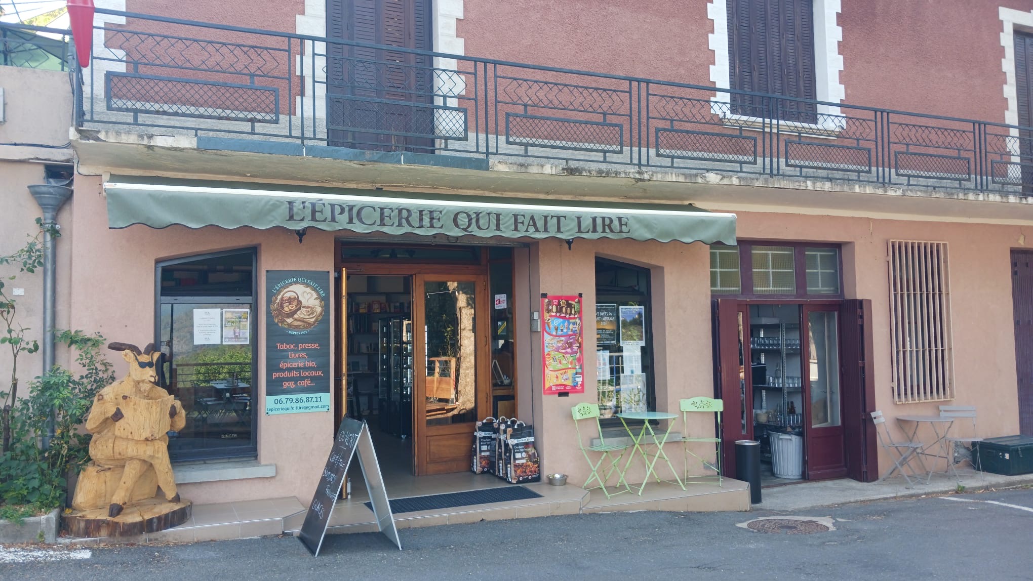 La devanture de L'Épicerie qui fait lire à Saint-Germain-de-Calberte : le store vert « L'Épicerie qui fait lire », la terrasse et la sculpture en bois d'un personnage qui lit