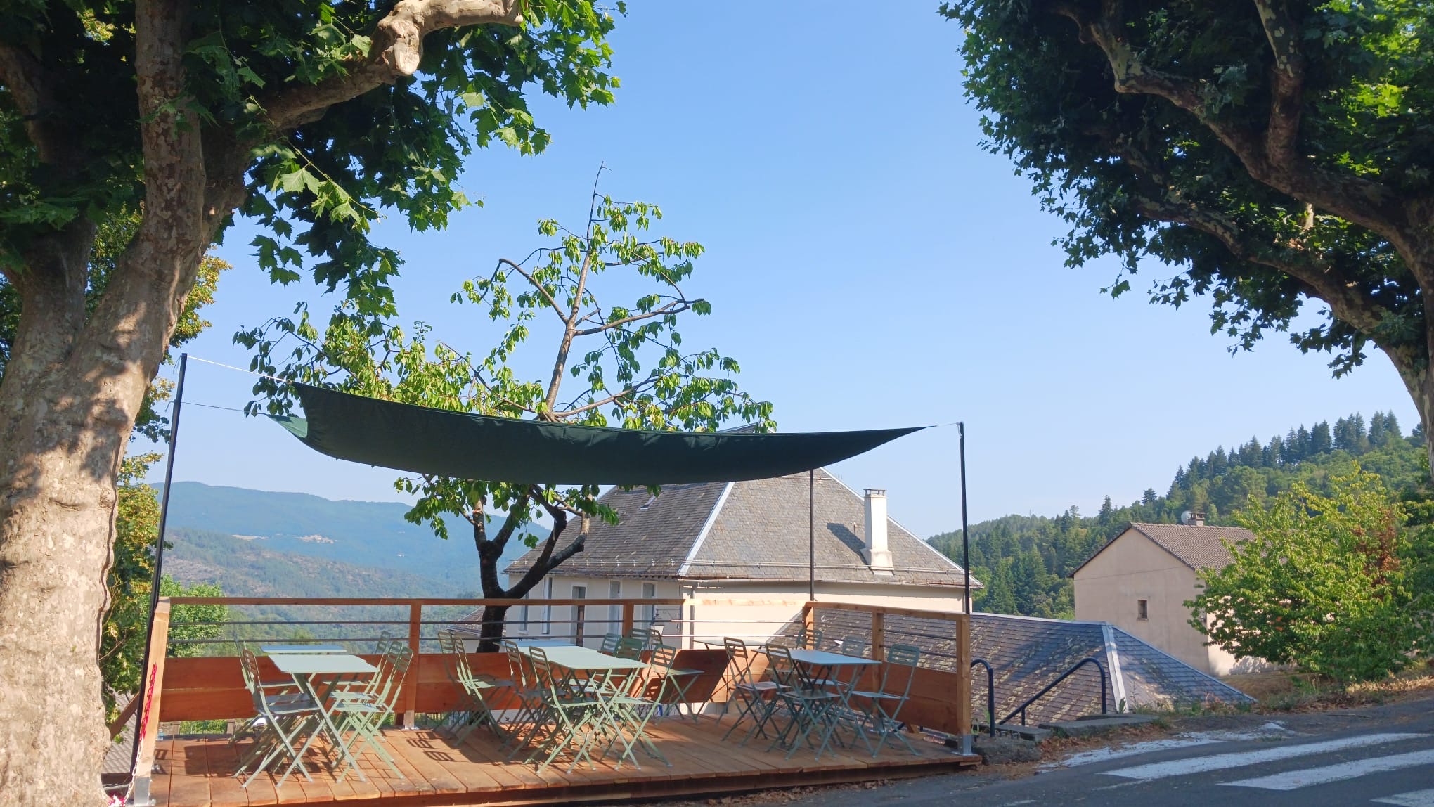 La terrasse de L'Épicerie qui fait lire : des tables et chaises vertes sous une voile d'ombrage, face aux montagnes des Cévennes